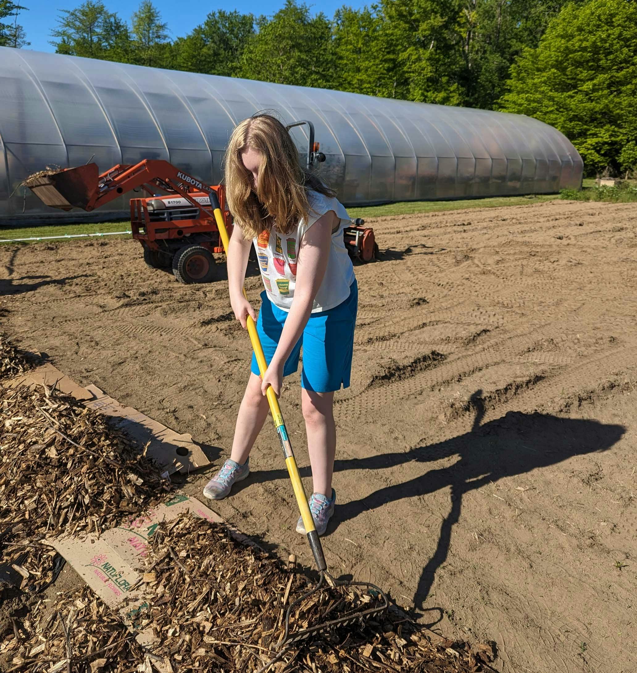 Riley is outside raking wood chips. A tractor and green house is behind her.