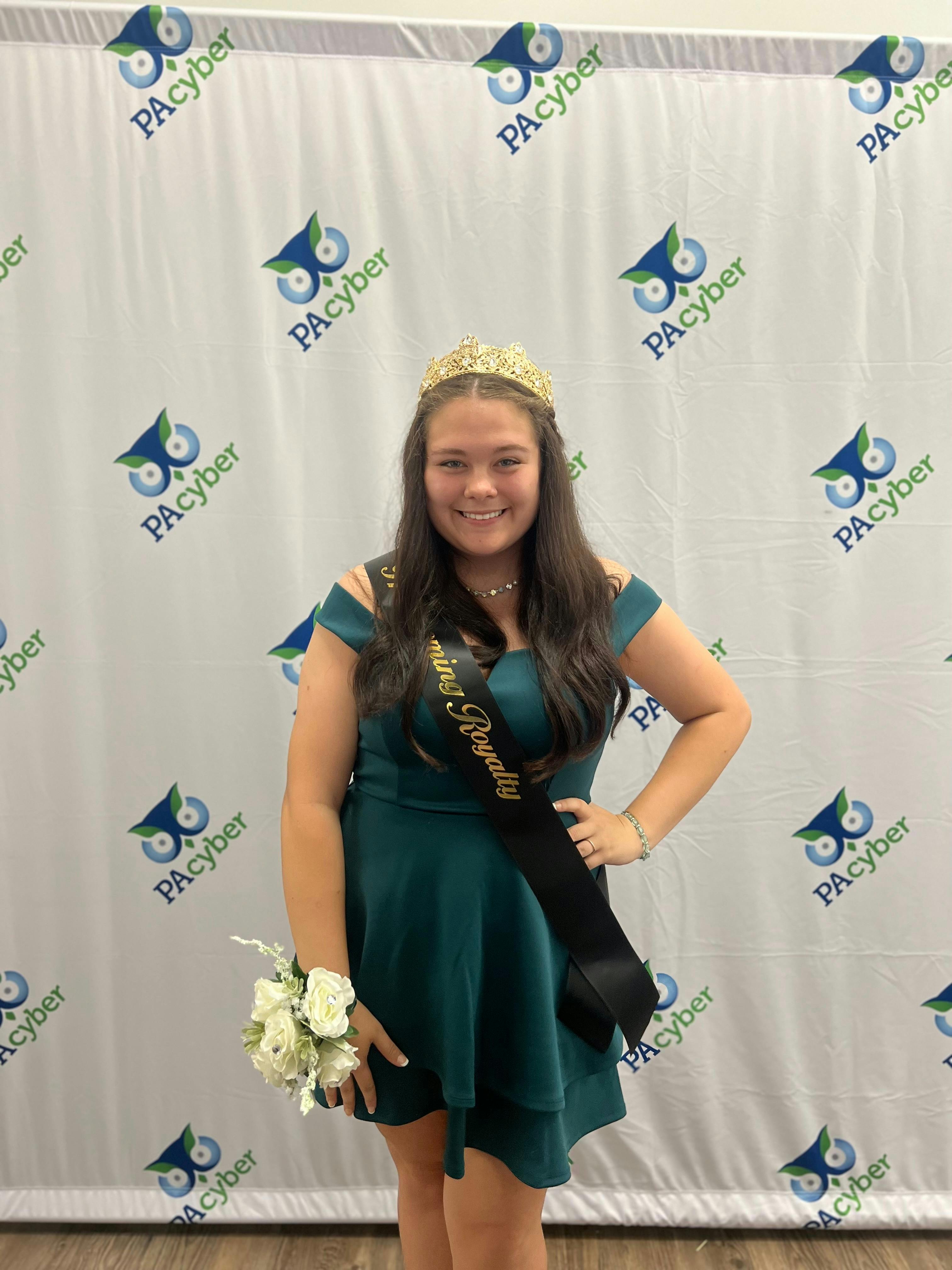 Kori wearing a prom queen crown, standing in front of a PA Cyber branded photo backdrop. She's wearing a short blue dress with a white flower corsage.