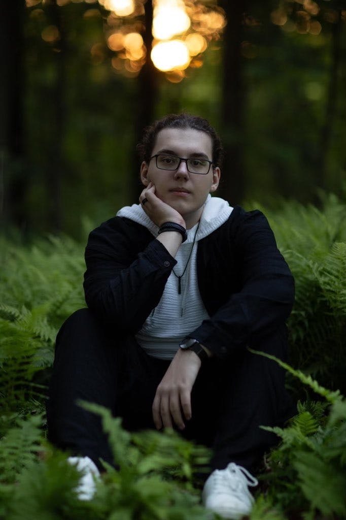 Landen smiling at camera, sitting on grass in the woods with his one arm resting under his chin and leg.