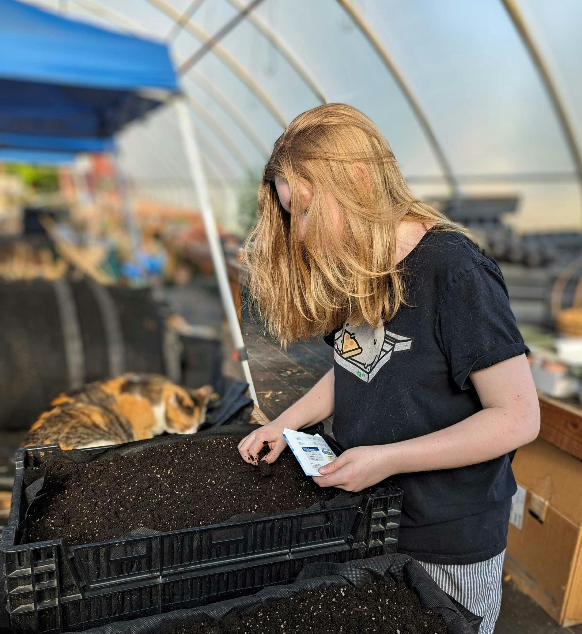 Riley is placing vegetable seeds in soil. She is inside a greenhouse. A calico cat lurks nearby.