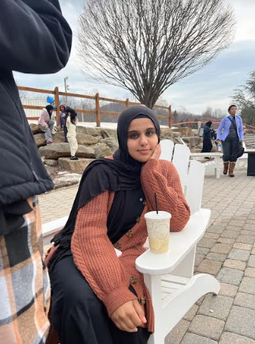 Reem is sitting on a white chair with her bubble tea sitting on the arm of her chair and her arm is resting under her chin.