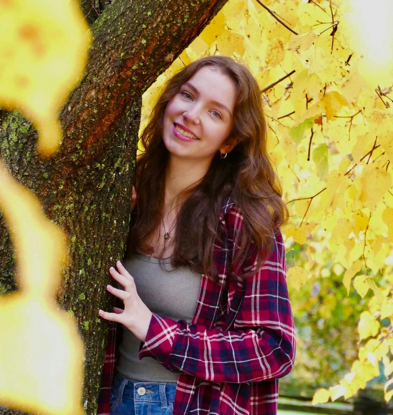 Young woman leaning against a tree. She is wearing a red and blue plaid shirt.