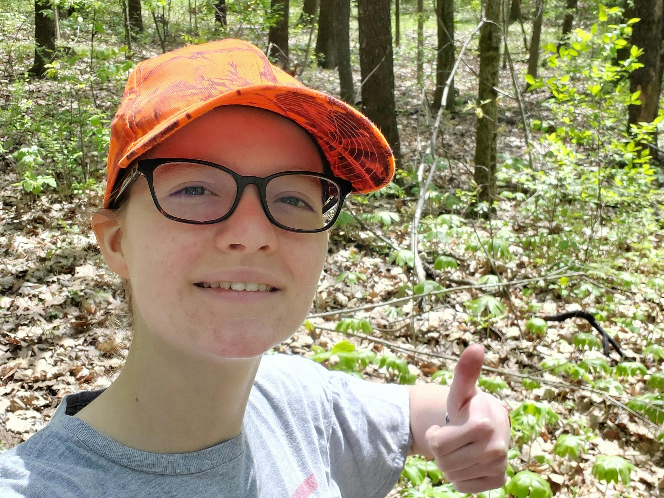 Jill wearing an orange baseball hat, taking a selfie with a thumbs up, with the woods in the background